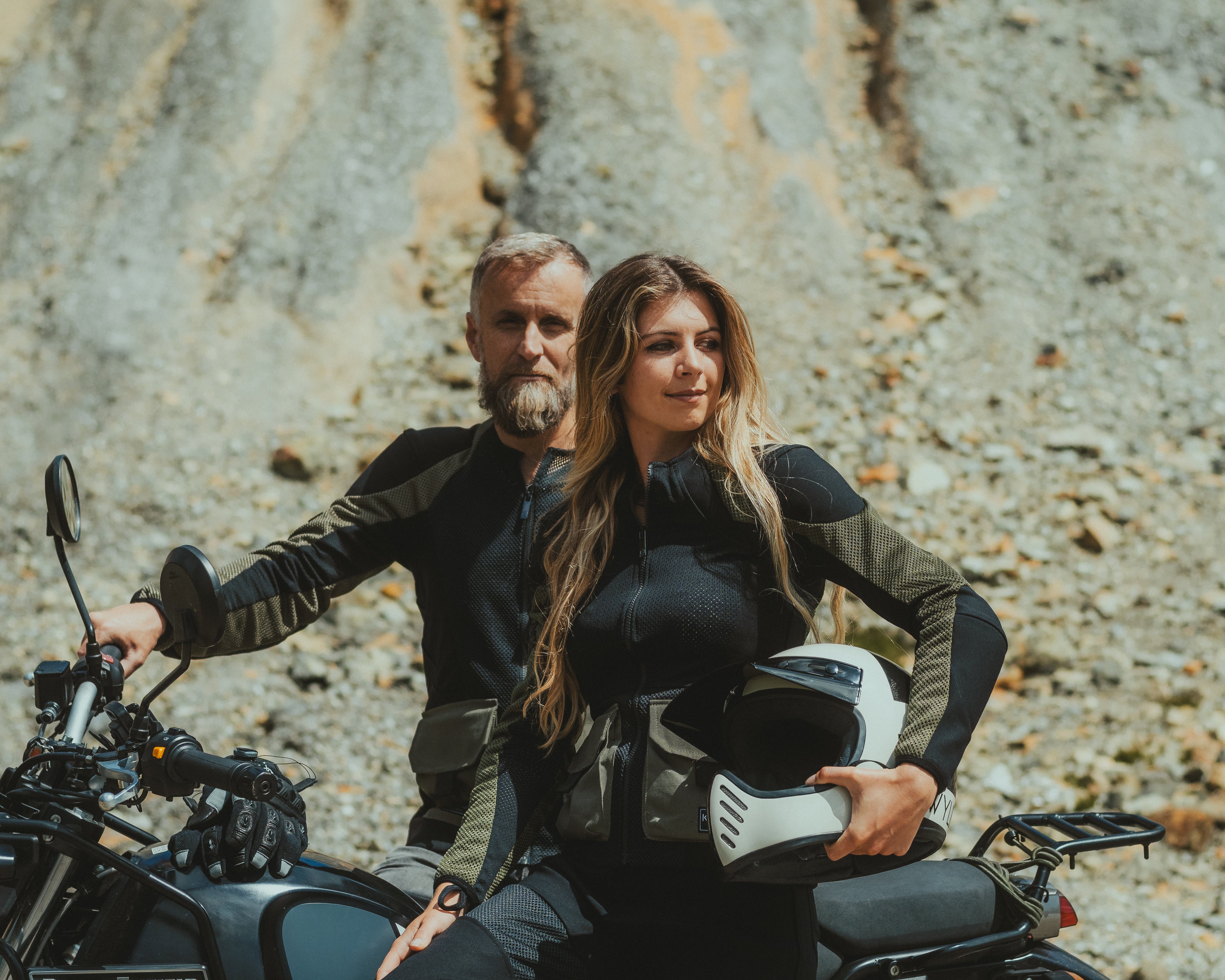 man and woman motorbike riders in protective motorbike gear by Knox. Sat and lent against a Royal Enfield motorcycle in Cornwall. Female rider holds a helmet by Atwyld.