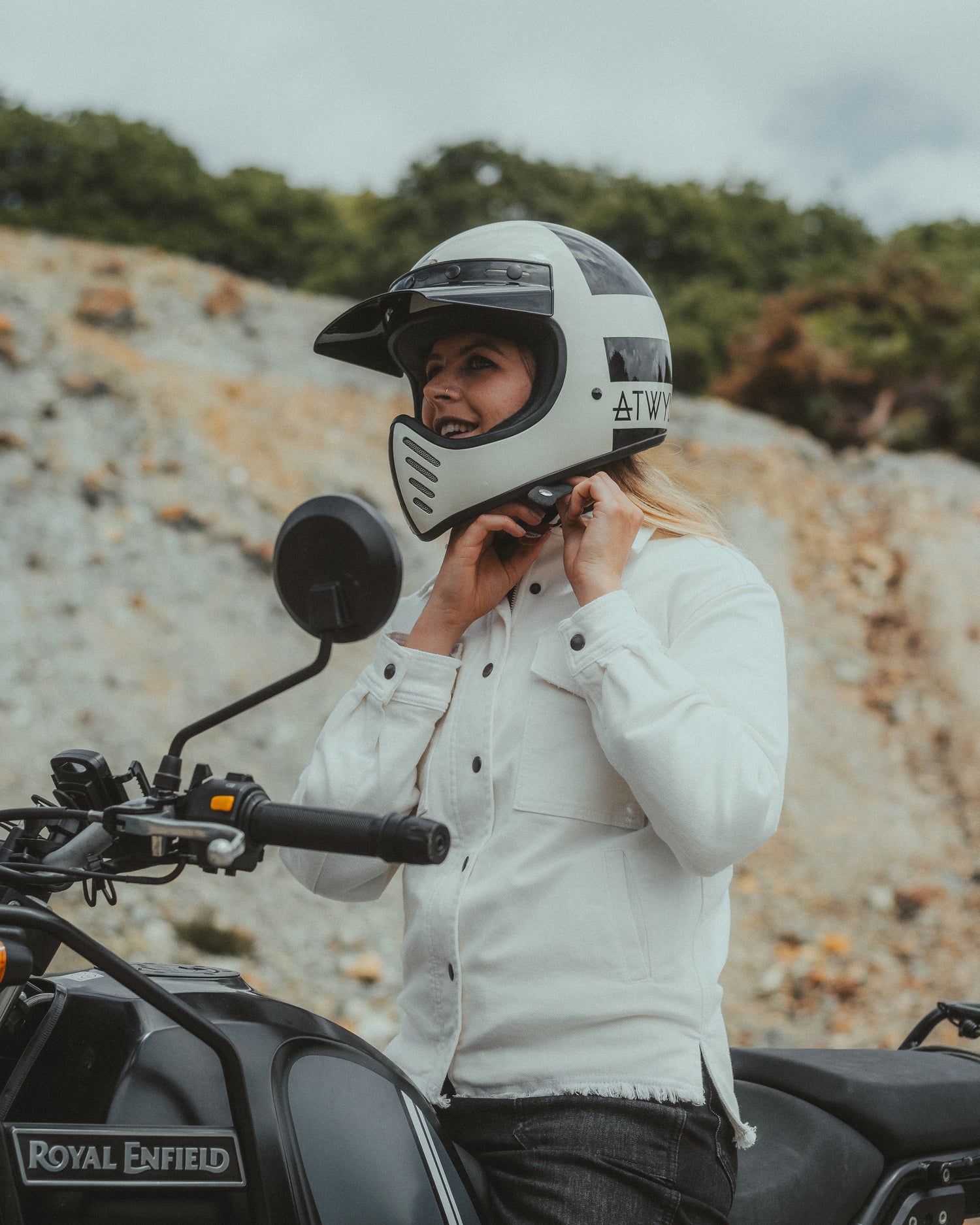 women motorbike rider sat on a Royal Enfield motorcycle wearing a protective white jacket and fastening a black and white helmet. Photo taken in Cornwall